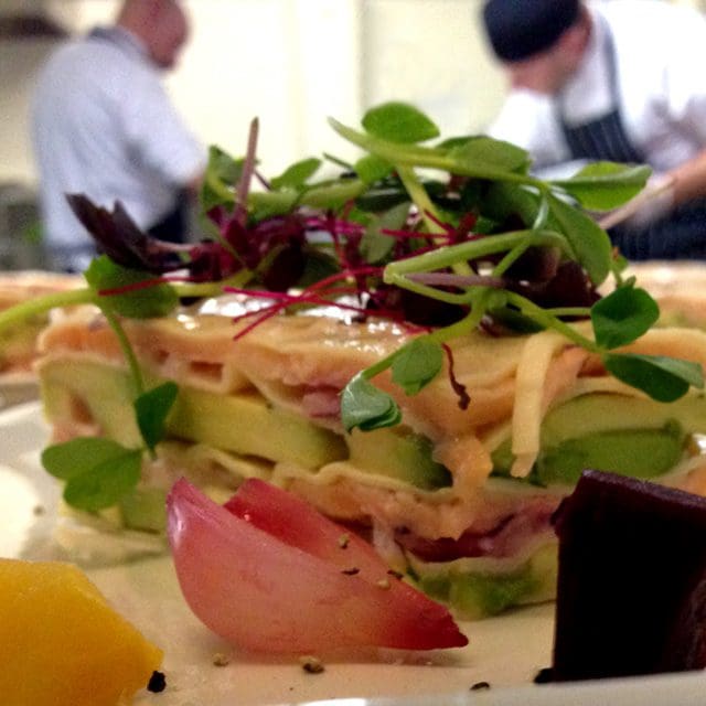 Contract catering kitchen, two chefs busy in the background with an avocado and trout lasagne topped with pea-shoots in the foreground, this leads to our web-form contact page